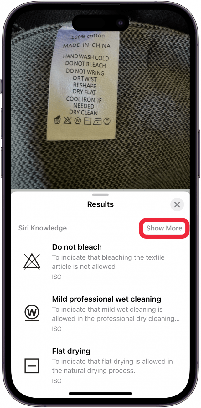 Die iPhone-Foto-App zeigt ein Foto eines Wäscheetiketts auf einem Pullover an, wobei das visuelle Suchmenü geöffnet ist, das verschiedene Wäschesymbole und ihre Bedeutung anzeigt. Die Schaltfläche "Mehr anzeigen" ist von einem roten Kasten umgeben.
